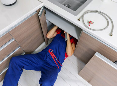 plumber working under domestic kitchen sink, repairing water pip